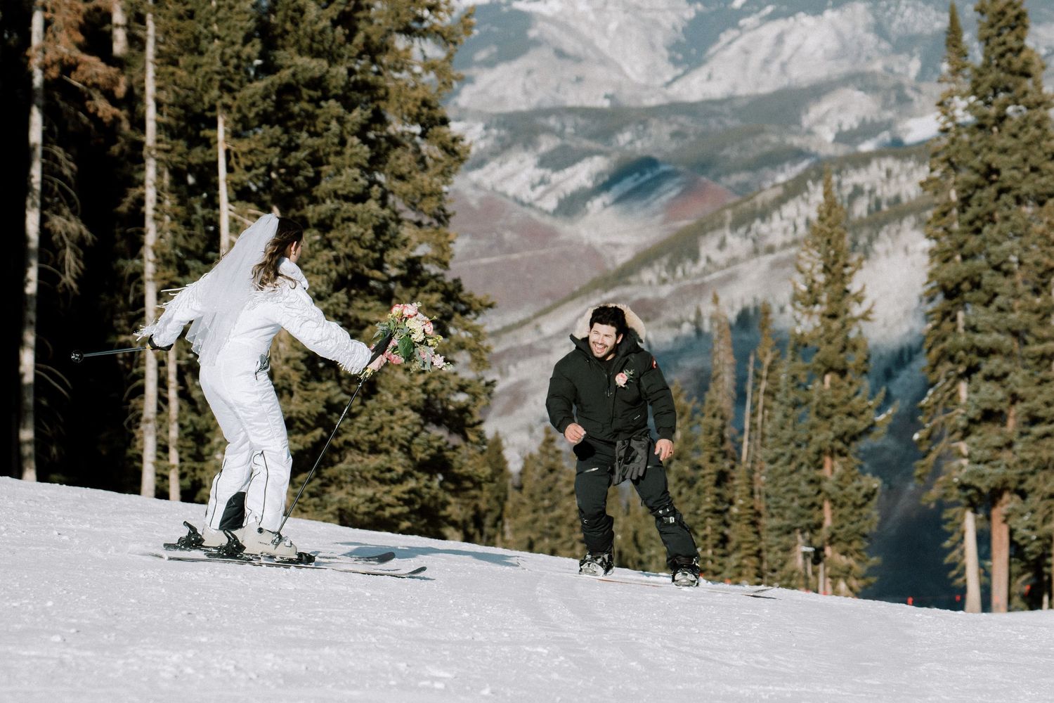 Two skiers glide down a snow-covered slope with evergreen trees and mountain peaks in the background.