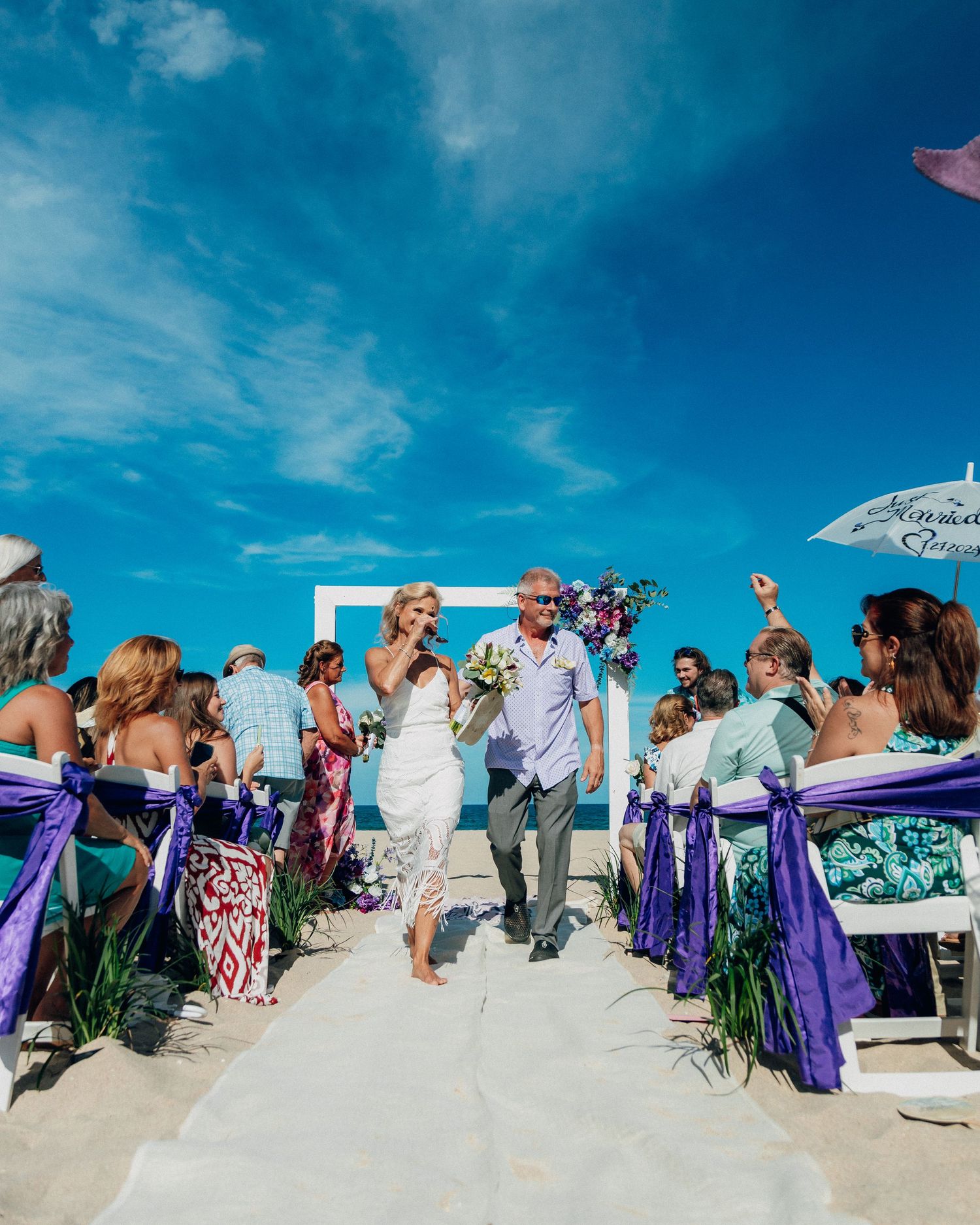 Beachside wedding ceremony with colorful decorations and guests under bright blue sky.