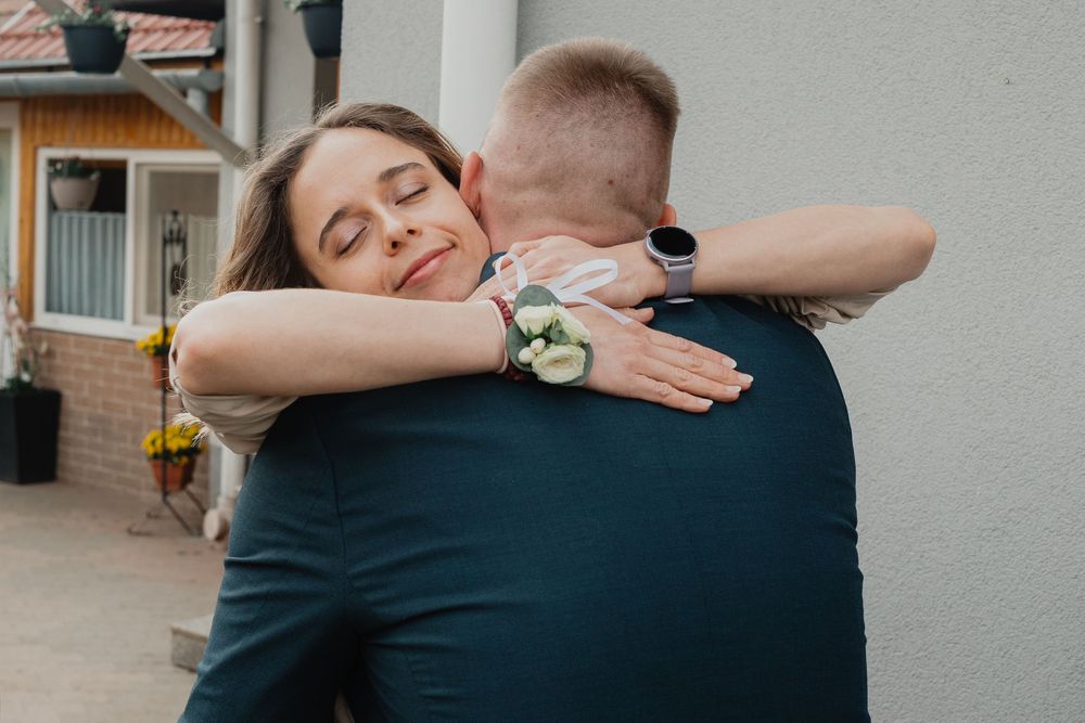 Two people embrace in a tender hug outside a house, with one wearing a navy blue shirt and white floral corsage.