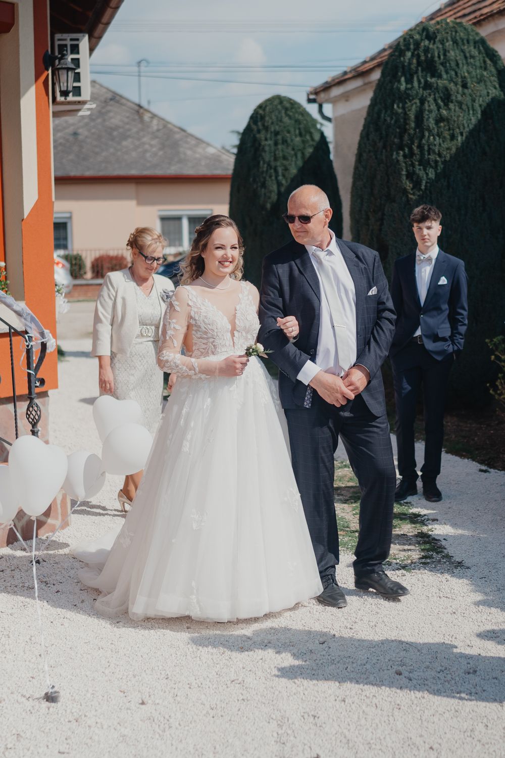 A bride in a white lace gown and veil stands outside on a gravel path with wedding party members in formal attire.