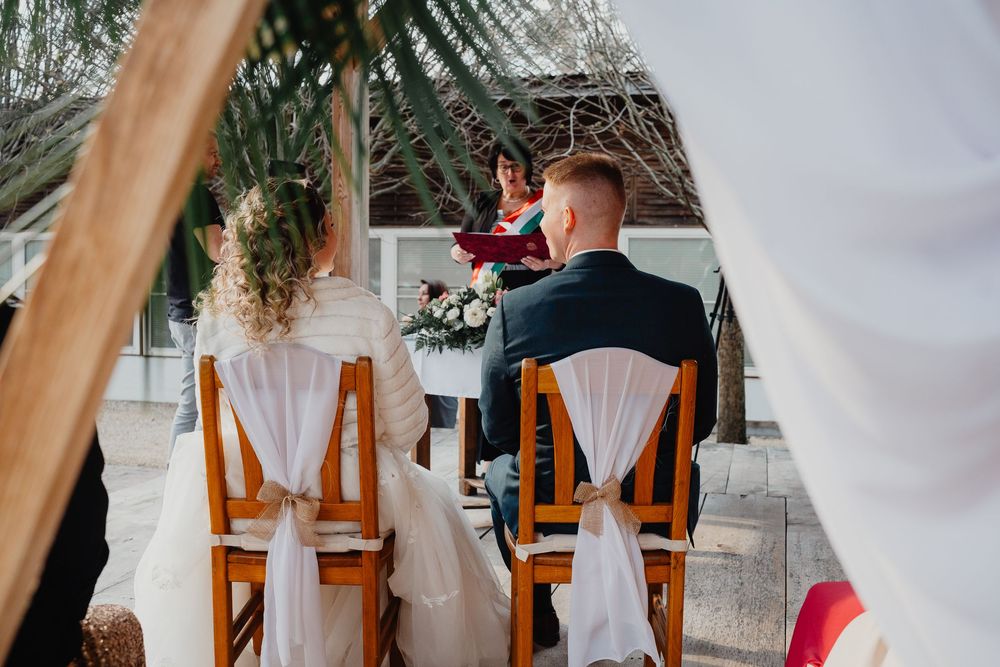 A couple sits in wooden chairs during an outdoor wedding ceremony under a draped arch and palm trees.