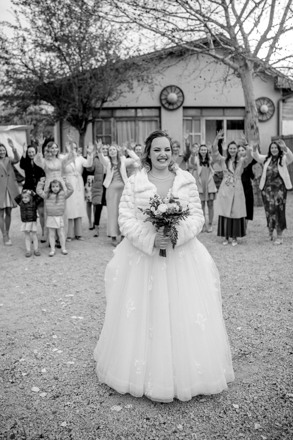 A black and white wedding photo showing a bride in a white gown holding a bouquet with guests celebrating in the background.