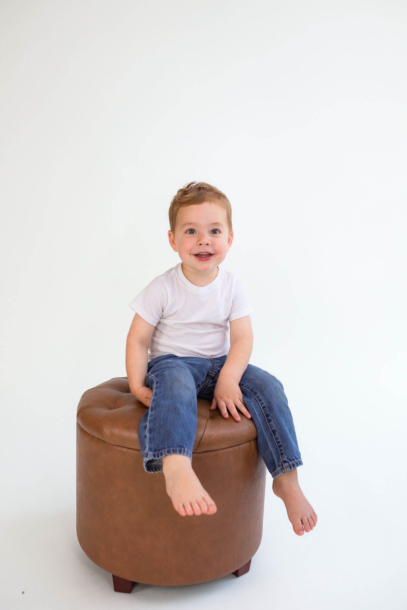 A child in a white t-shirt and jeans sits on a round brown leather ottoman against a white background.