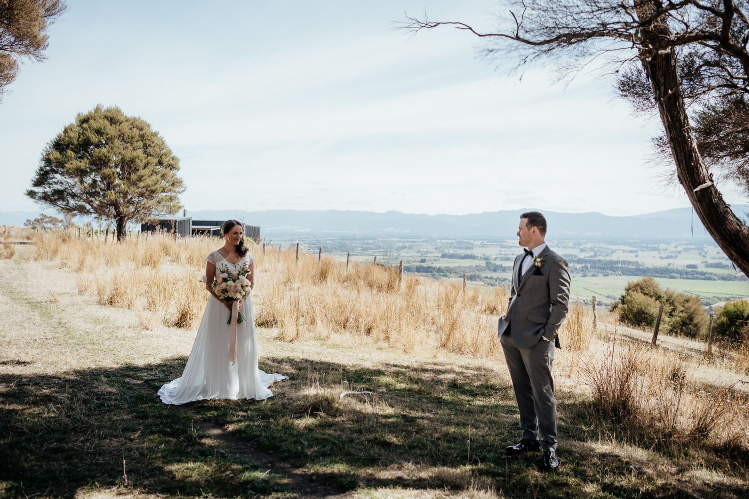 A couple stands apart on a hilltop with sweeping mountain views and golden grass on a sunny day at Whitimanuka by August and Vine