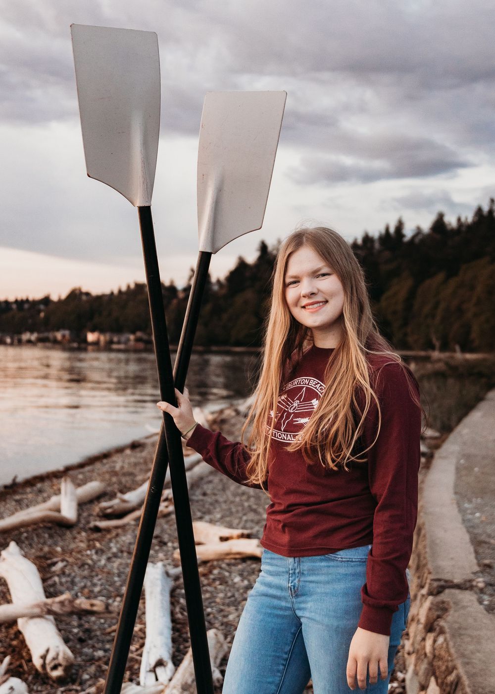 Long-haired person in burgundy sweater and jeans holds kayak paddles while standing by rocky shoreline at sunset.