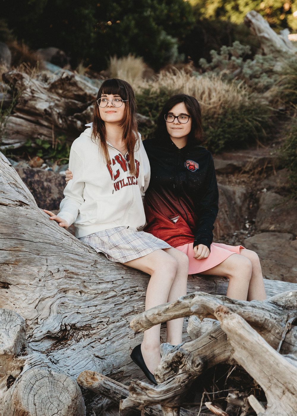 Two students sit on driftwood logs in an outdoor setting wearing casual attire with glasses and smiles.