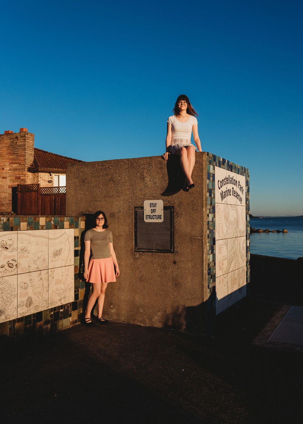 Two people pose at sunset near an oceanfront wall with brick structures and a blue sky backdrop.