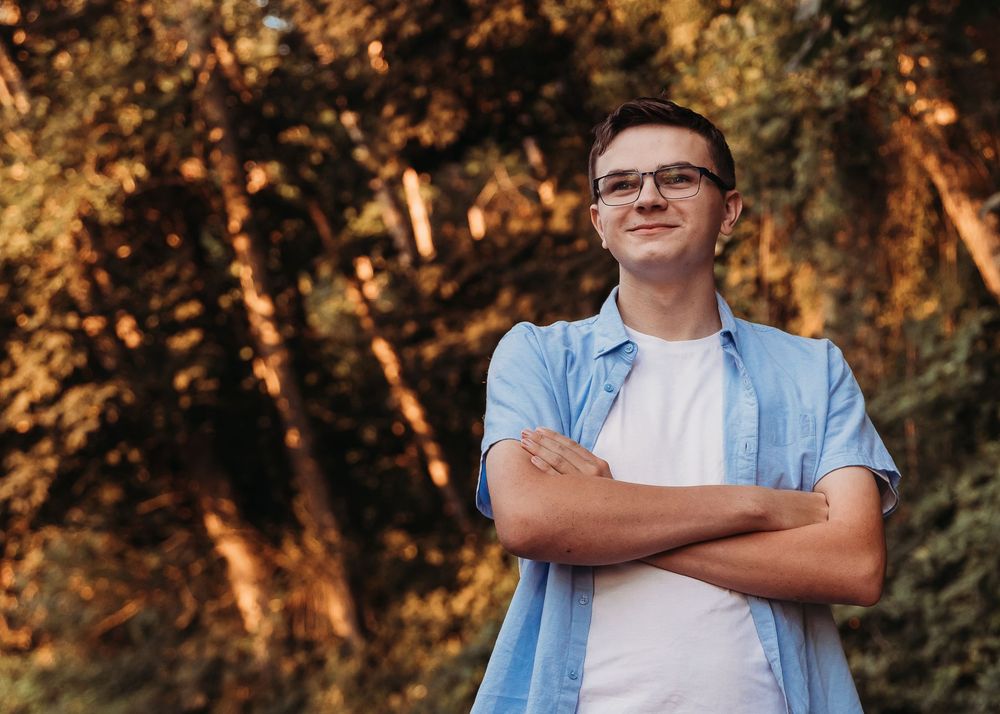 Young person in glasses and blue button-down shirt stands with crossed arms in front of sunlit autumn trees.