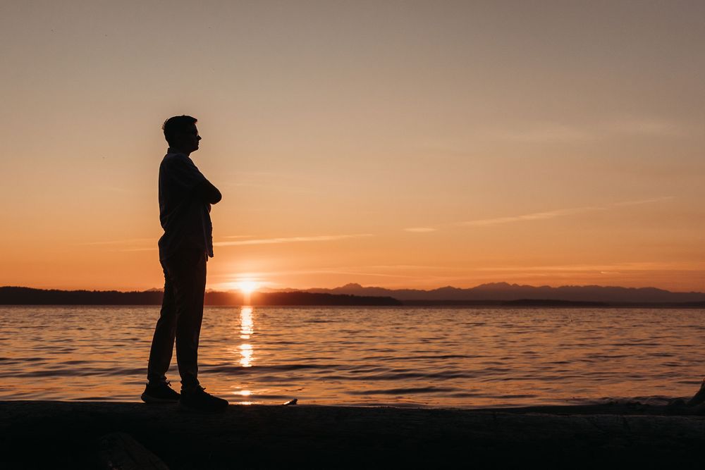 A silhouetted figure stands at the water's edge during a vibrant orange sunset over a tranquil lake.