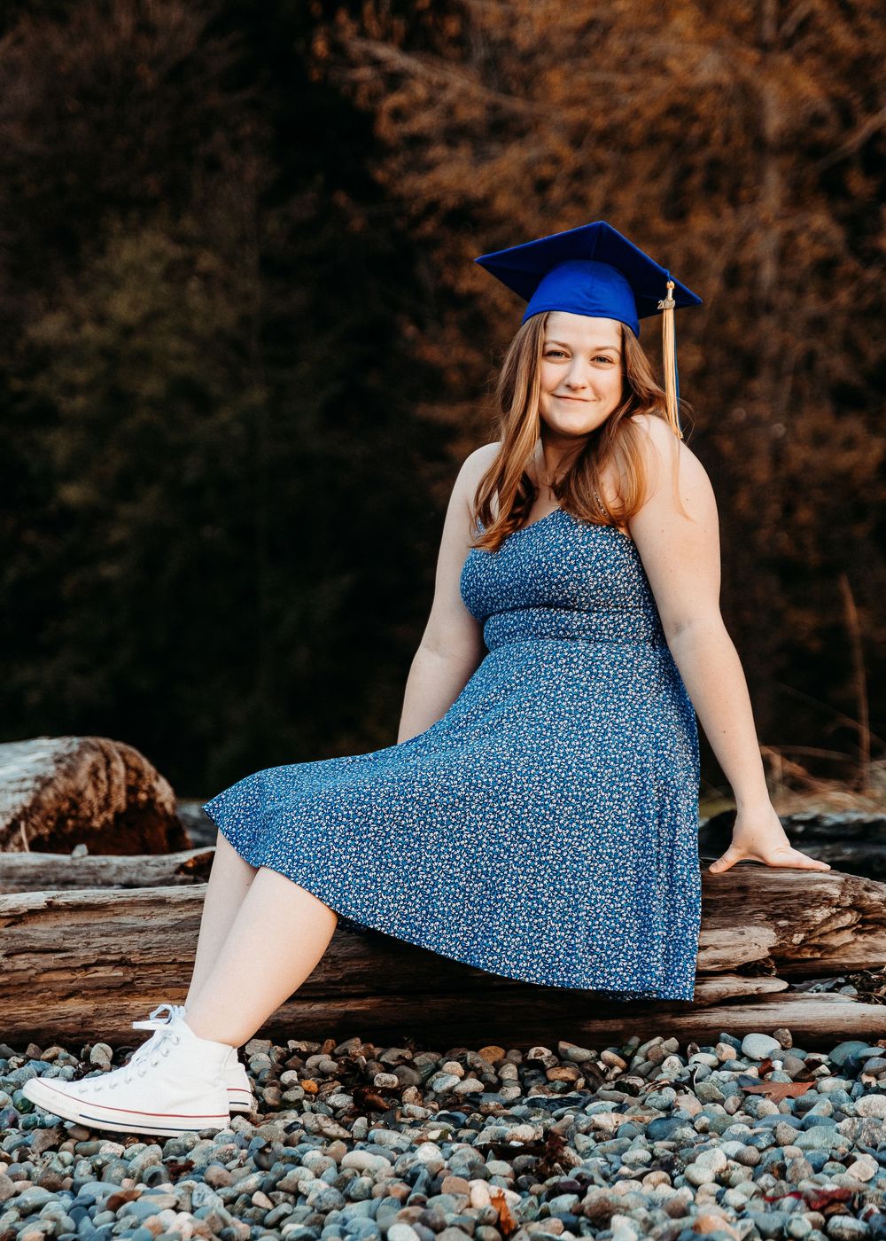 A graduate in blue dress and blue cap sits on driftwood at a rocky beach during autumn sunset.