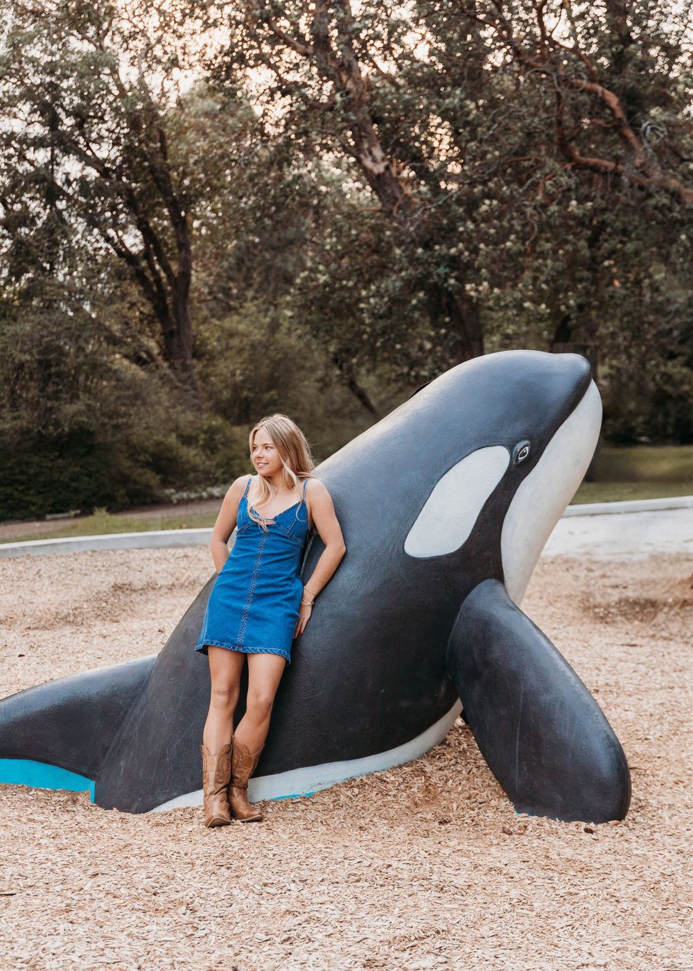 A figure in a blue dress poses next to a large decorative orca whale statue on a gravel surface near trees.