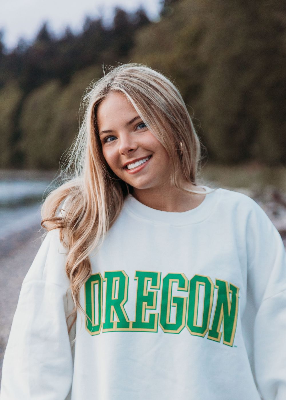 Blonde wearing a white sweatshirt with green Oregon university text at beach with forest background.