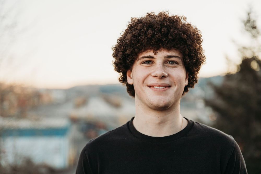 Portrait of a person with curly dark hair and black t-shirt smiling outdoors during sunset.