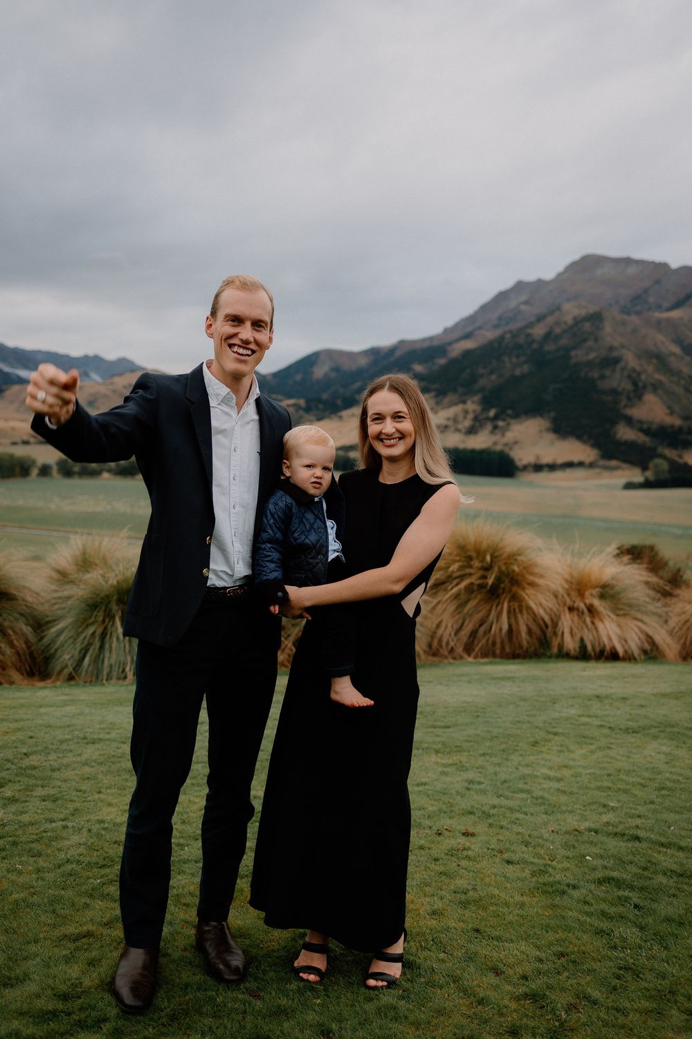Family of three poses together on scenic mountain lawn with moody cloudy sky backdrop.
