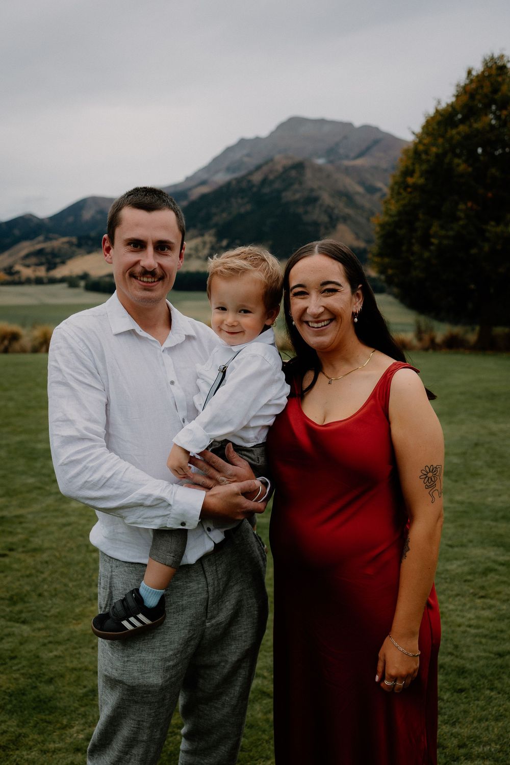 A small family stands together on grassy field with scenic mountains in the background during overcast weather.