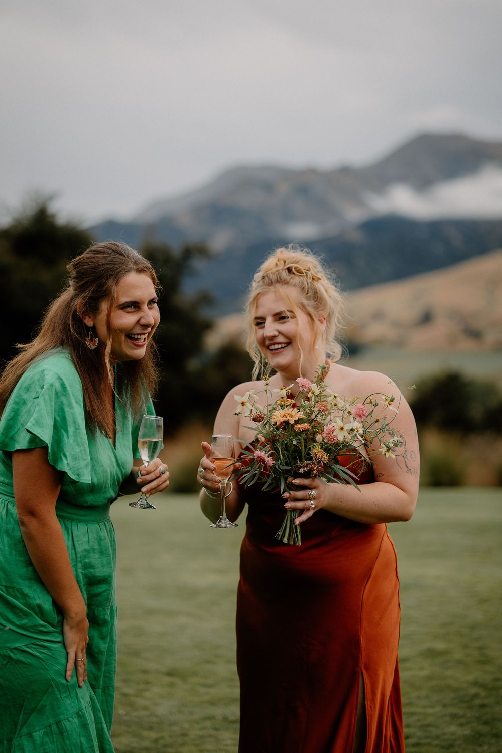 Two women share a joyful moment in a field with mountains, one in green dress holding wine, other with wildflower bouquet.