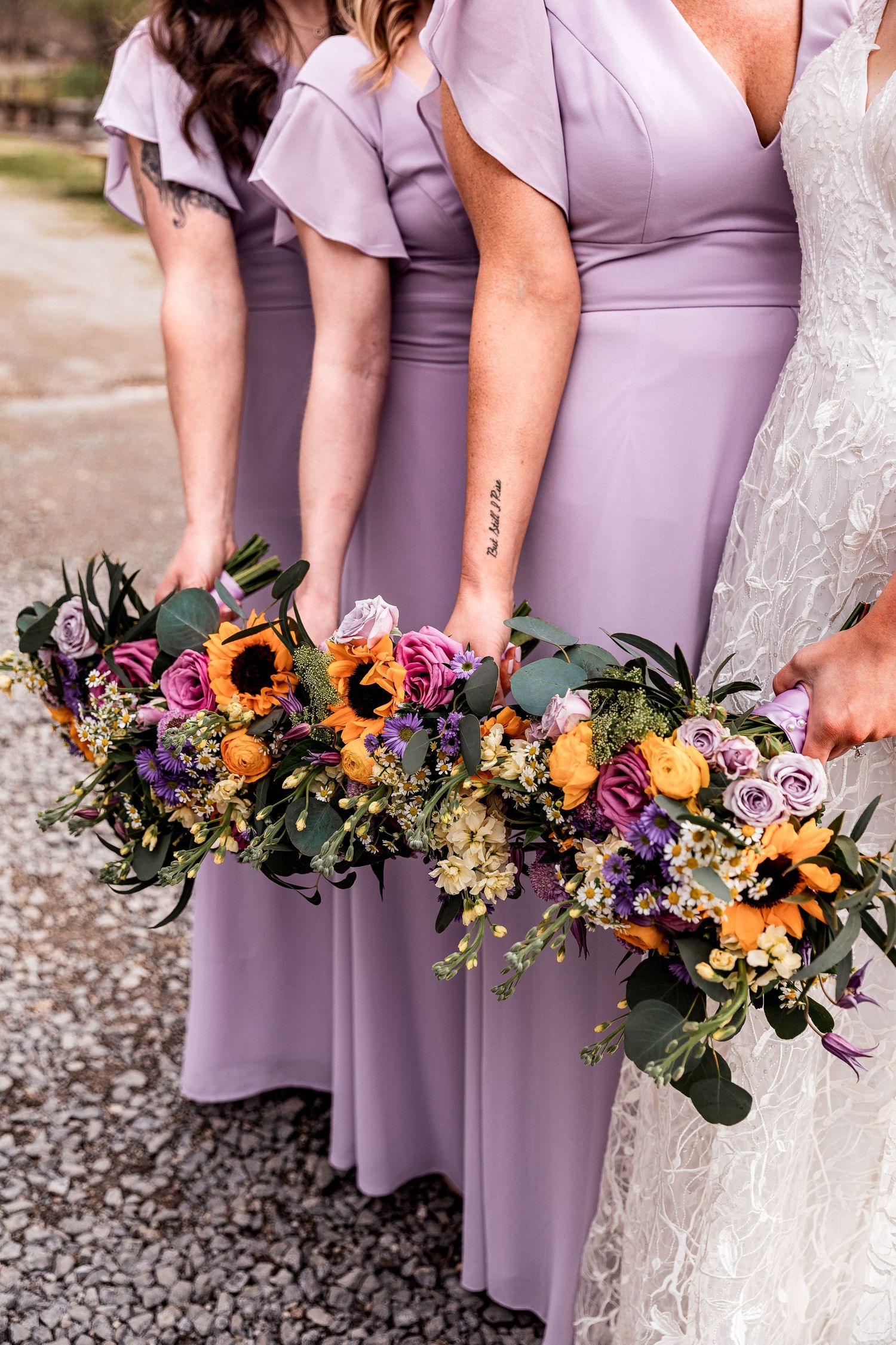 Line of bridesmaids in lavender dresses holding vibrant bouquets with sunflowers, roses and purple flowers at outdoor wedding.