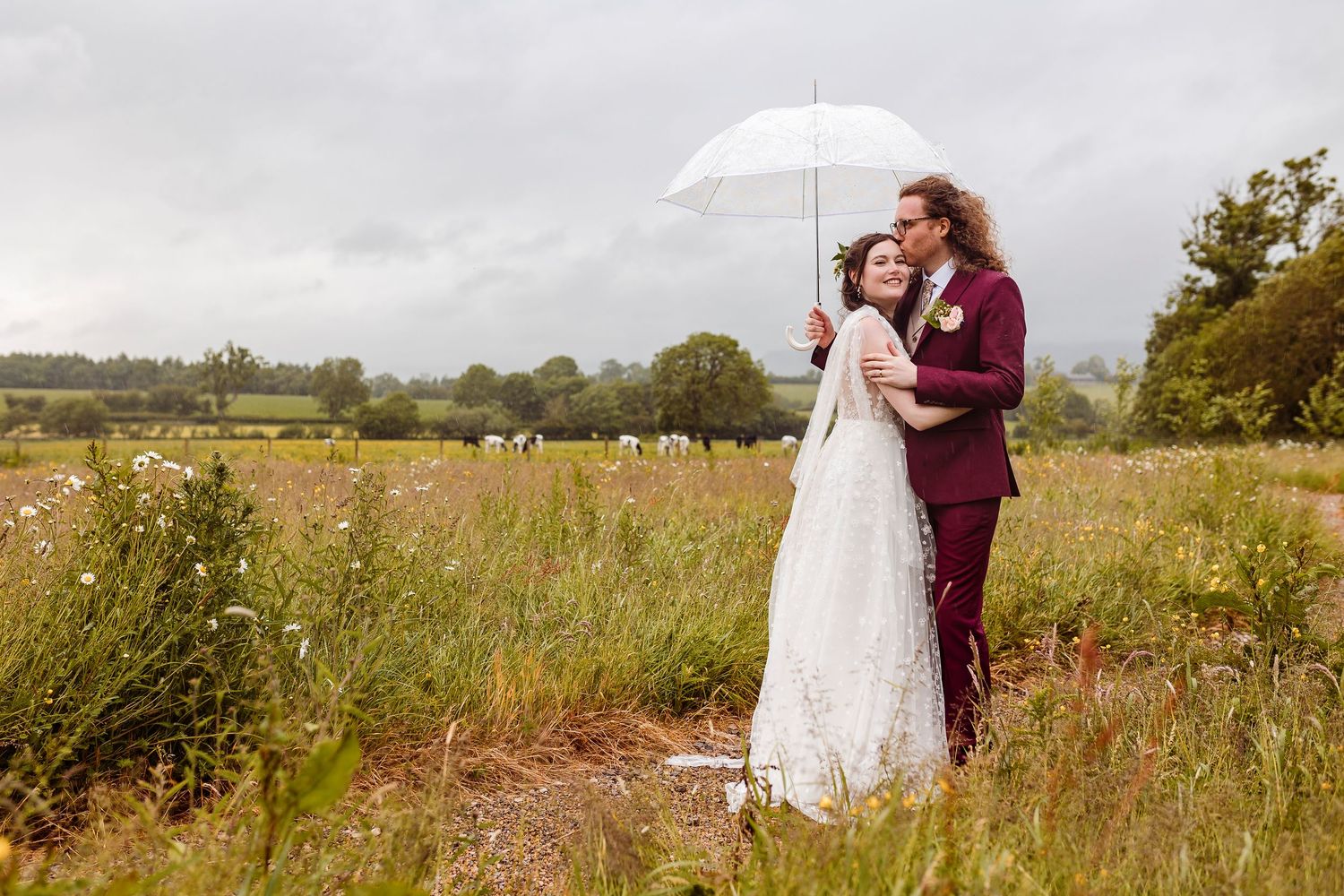 A couple shares a romantic moment under a white umbrella in a wildflower meadow on their rainy wedding day.
