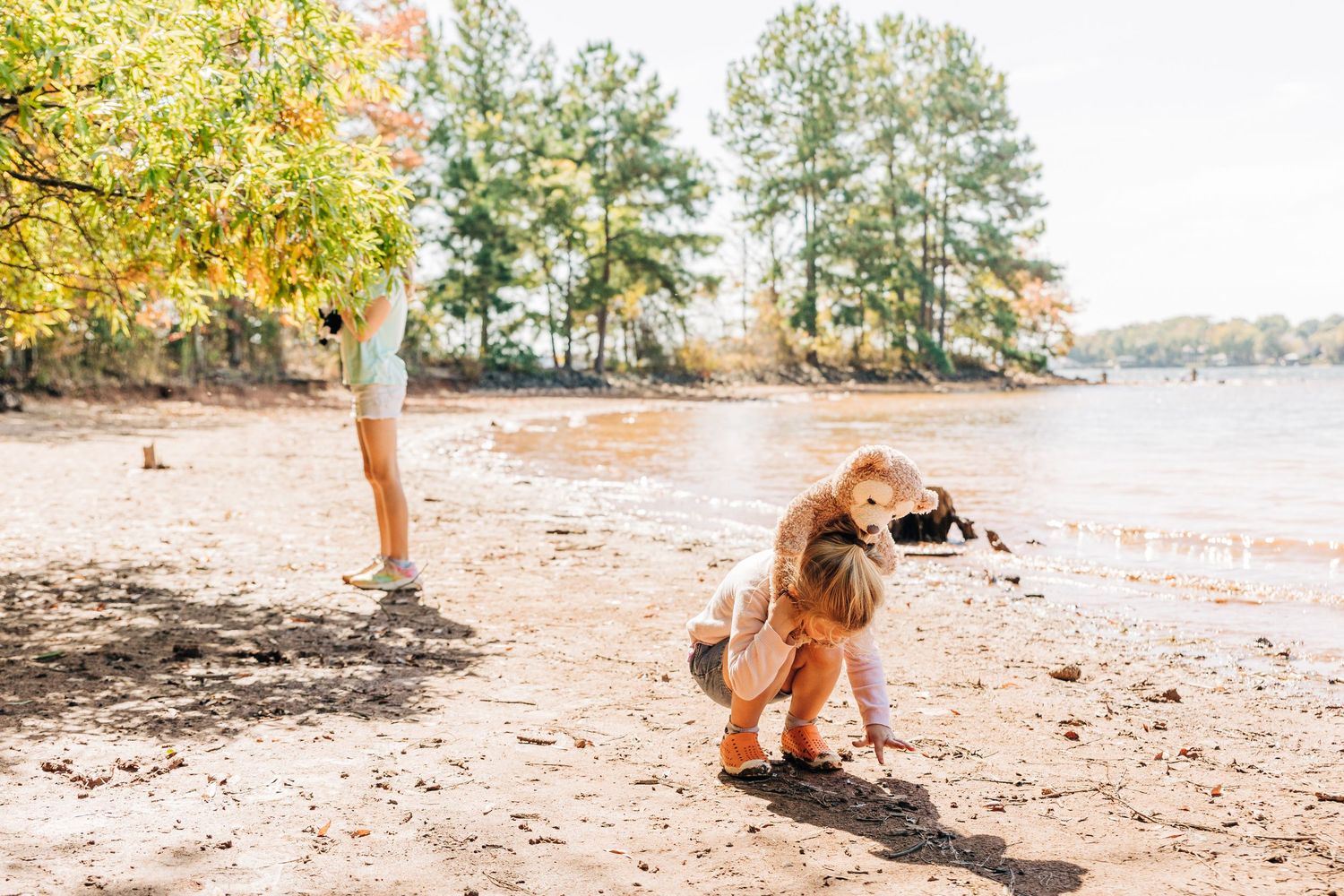 Jetton Park beach image of young girl digging in sandy soil near pine trees and water with teddy bear on her shoulders