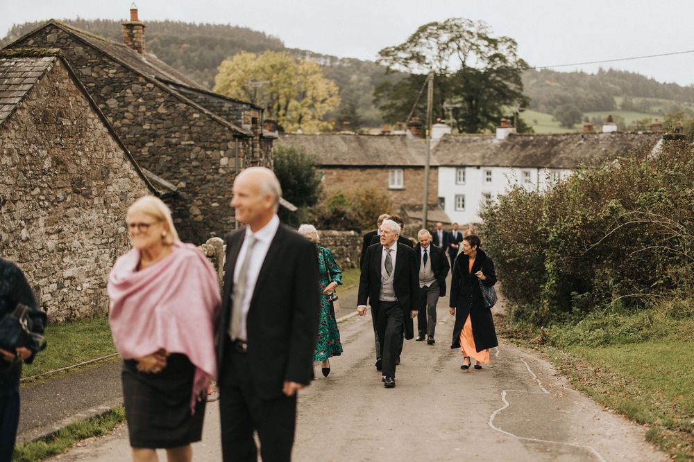 Wedding guests walk along a country lane beside stone buildings and trees in a rural English village setting.