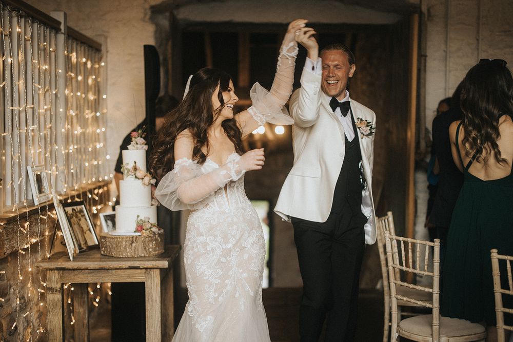 A couple in elegant wedding attire dances joyfully at their indoor reception with twinkle lights.