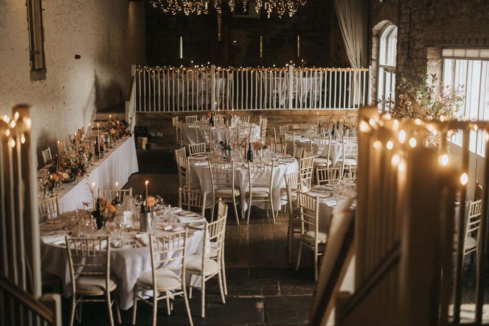 Elegant wedding reception venue with candlelit round tables, white chairs and a rustic balcony in warm ambient lighting.