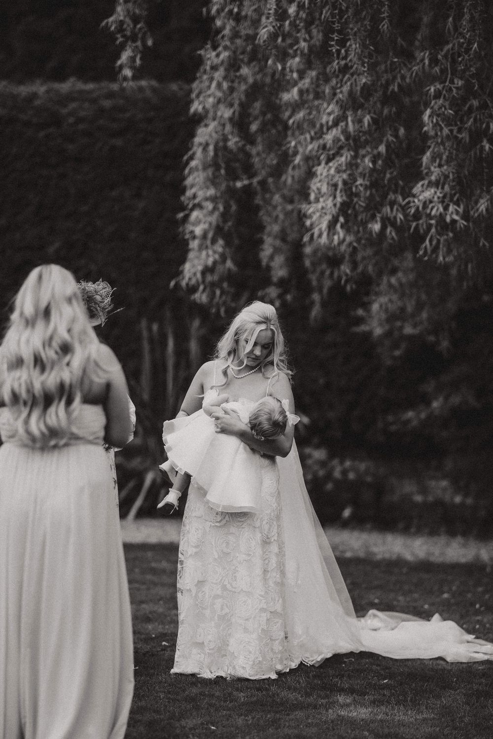 Two women in light-colored dresses walk together through a garden setting in black and white photography.