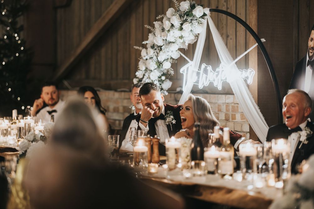 Wedding reception guests share joyful laughs at an elegant rustic barn venue decorated with white florals and candles.