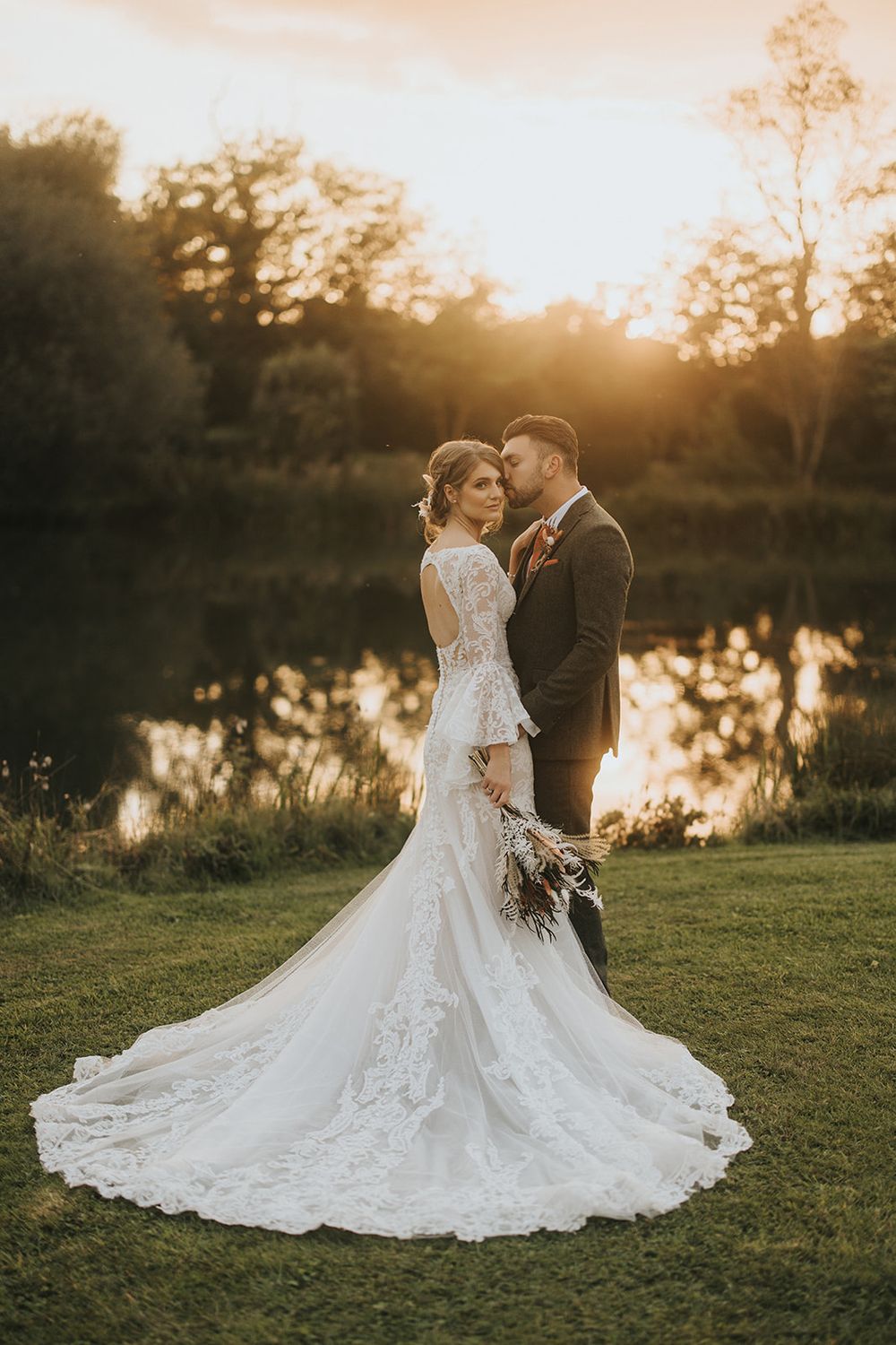 A romantic wedding portrait at sunset by a lake with the bride wearing a long lace train dress and holding a bouquet.