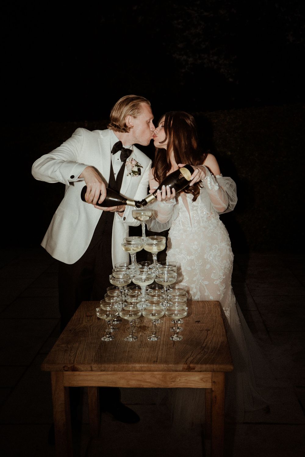 Newlyweds pour champagne into an elegant tower of glasses on a rustic wooden table during a dark outdoor reception at Askham Hall.