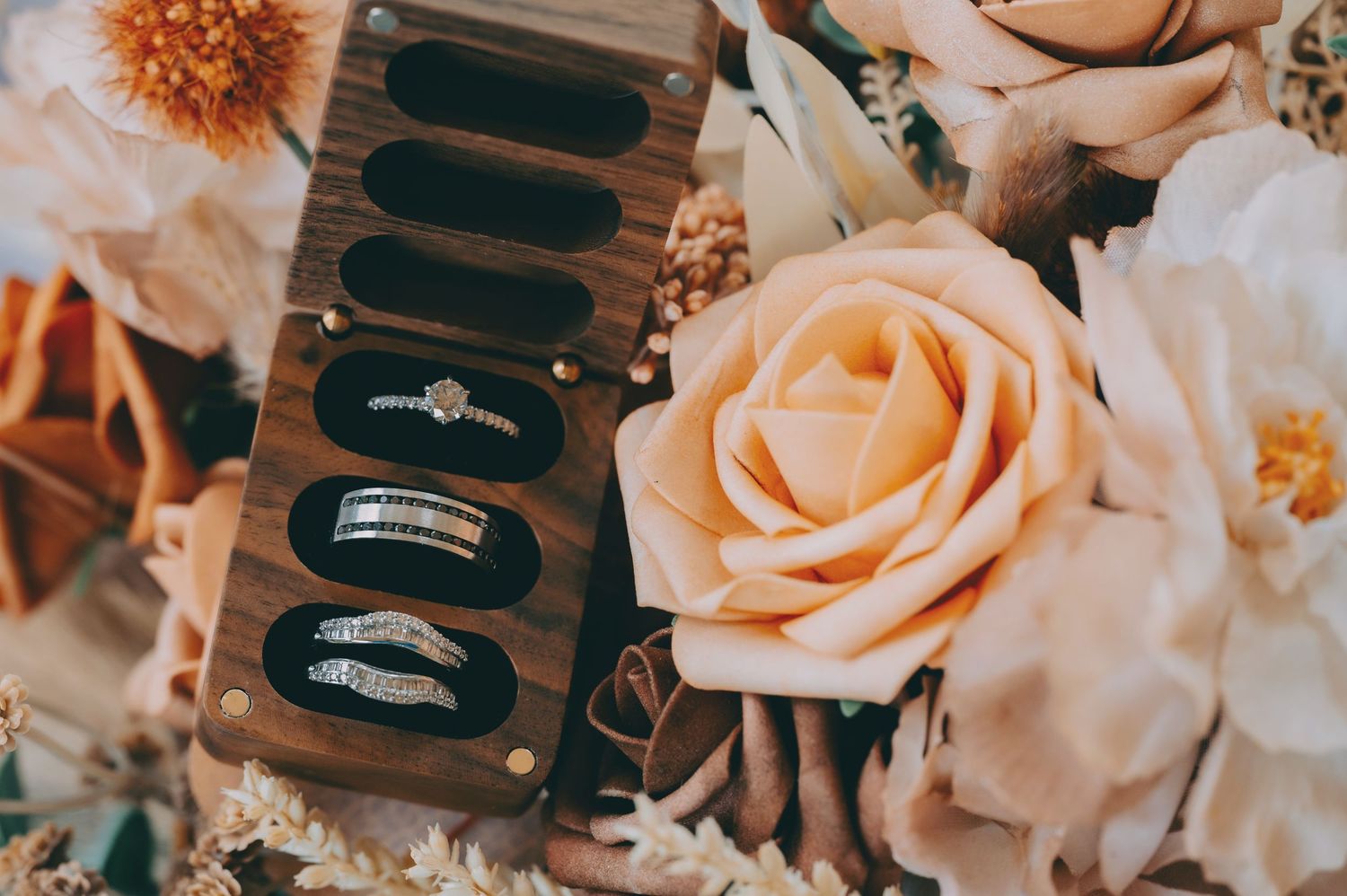 Wedding rings displayed on dark velvet cushions alongside soft peach roses and floral arrangements.