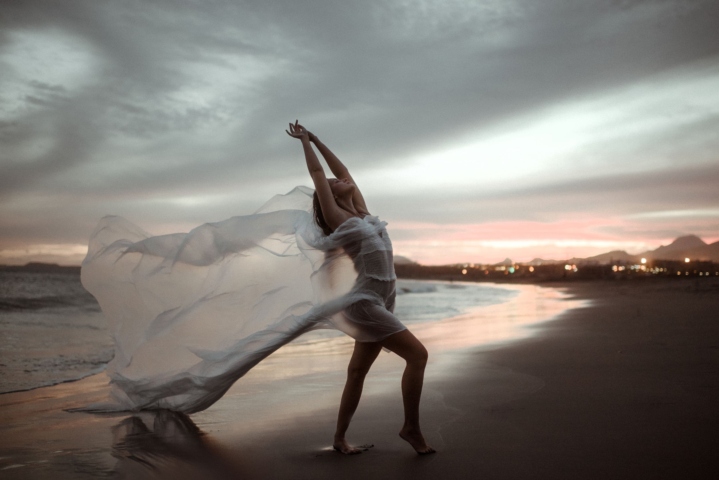 A young woman wearing a white body performs an elegant movement on La Tejita beach at sunset against dramatic, cloudy skies.