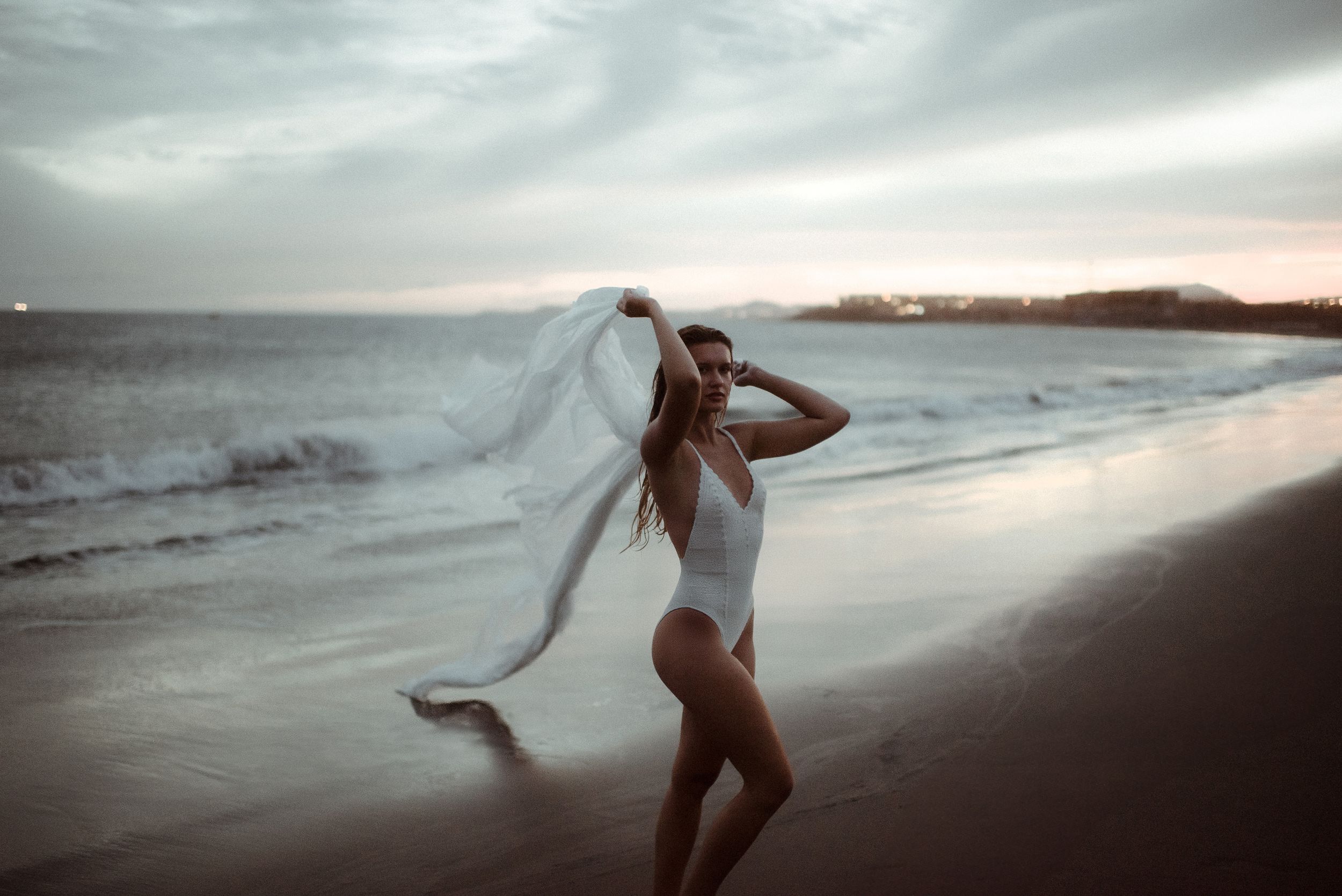 A woman in a white swimsuit walking at La Tejita beach at sunset with flowing fabric in the wind. Portrait photoshoot in Tenerife.