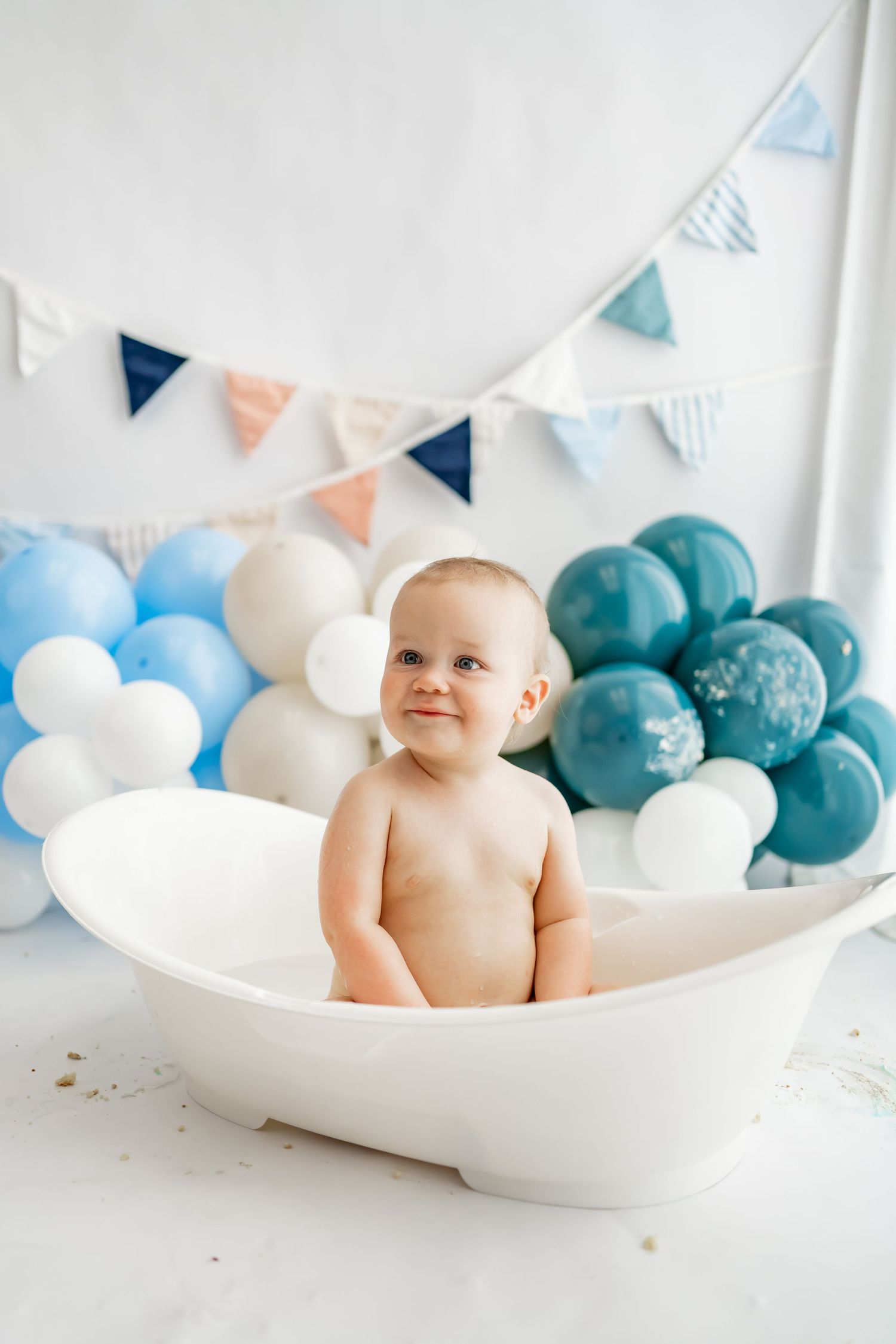 Decorative baby photo setup with white bathtub, blue and white balloons, and pastel bunting flags against white backdrop.