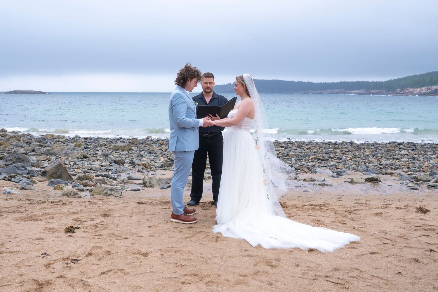 A romantic beach wedding ceremony takes place on a sandy shoreline with cloudy skies and ocean waves in the background.