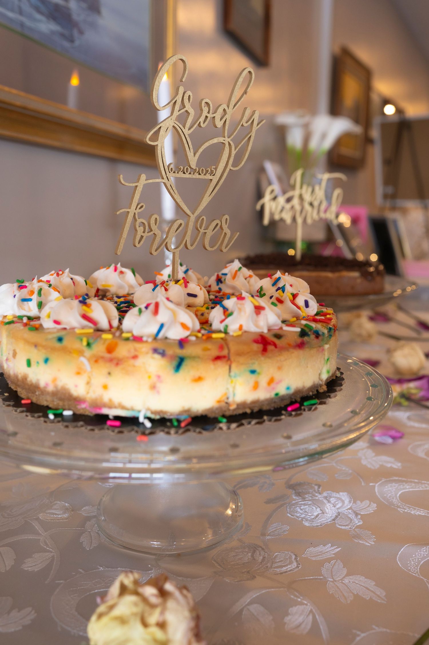 Wedding cake decorated with colorful sprinkles and gold script topper displayed on a clear glass cake stand.