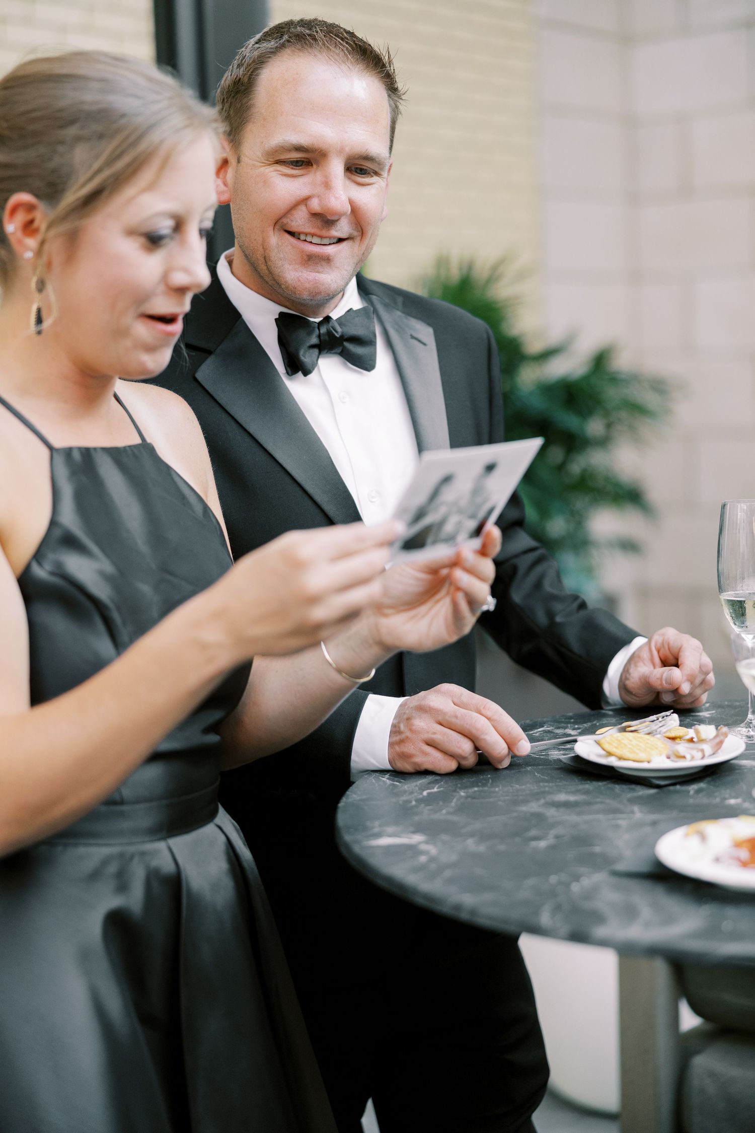 Formal event scene showing a couple in black tie attire looking at phones while enjoying appetizers at a table.