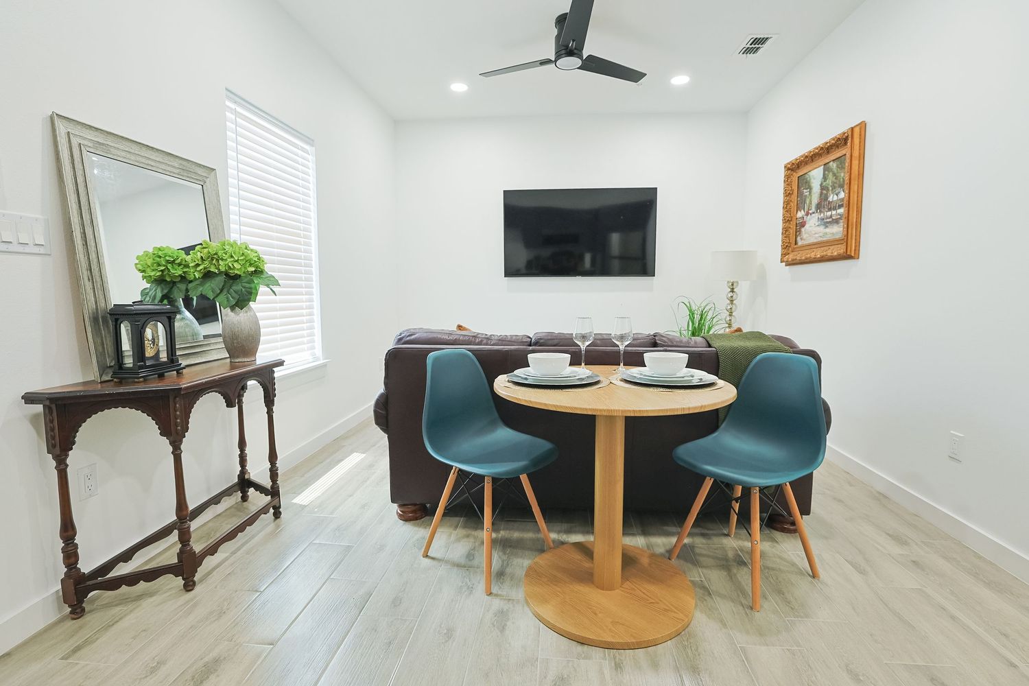 Modern living room corner with round dining table, teal chairs, gray sofa, and wall-mounted TV in bright white space.