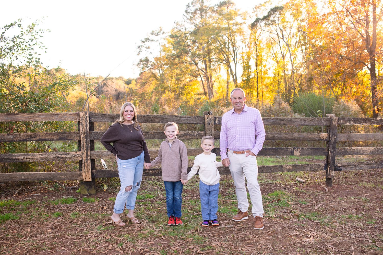 A family of four stands together in front of a rustic wooden fence during golden hour in an autumn setting at Sugg Farm