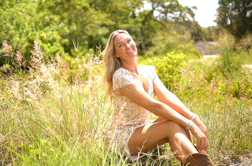 A person in a lace dress sits in a sunlit meadow with tall grass and wildflowers on a warm summer day.