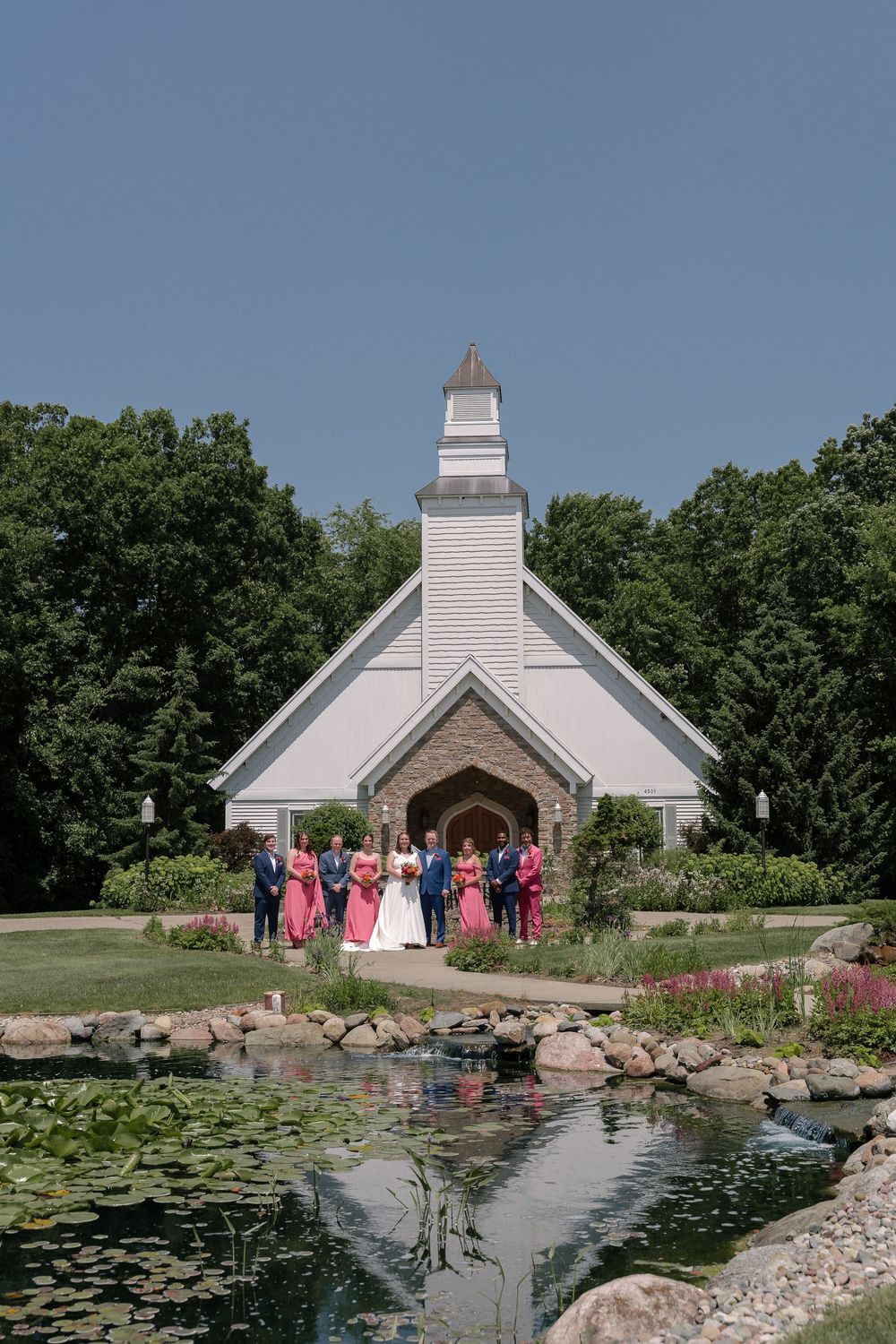 A picturesque white chapel with a steeple stands beside a lily pond during a summer wedding ceremony.