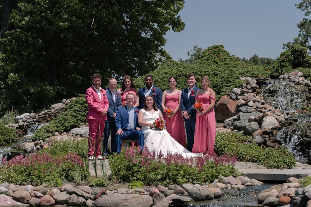 A wedding party stands in a scenic outdoor garden setting with lush greenery and stone features in the background.