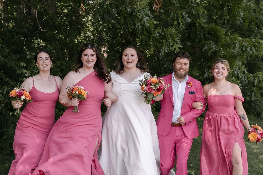 Wedding party poses together in coordinating pink and white attire near trees at outdoor celebration.
