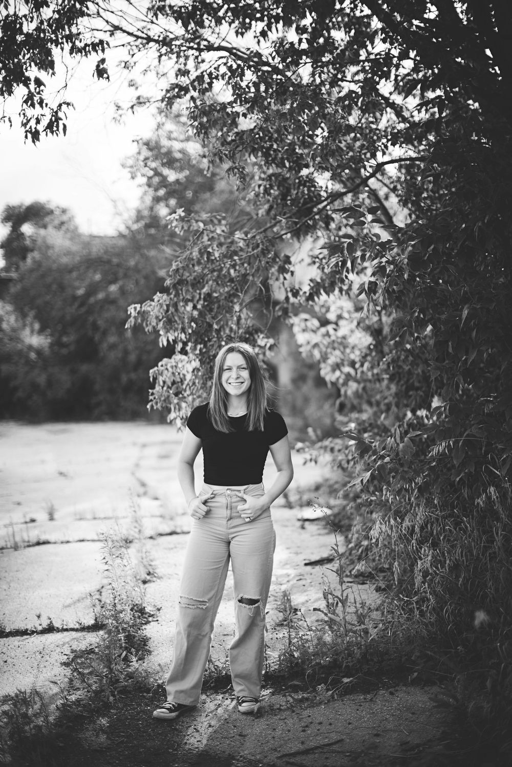 Black and white outdoor portrait of someone standing on a path under overhanging tree branches at sunset.