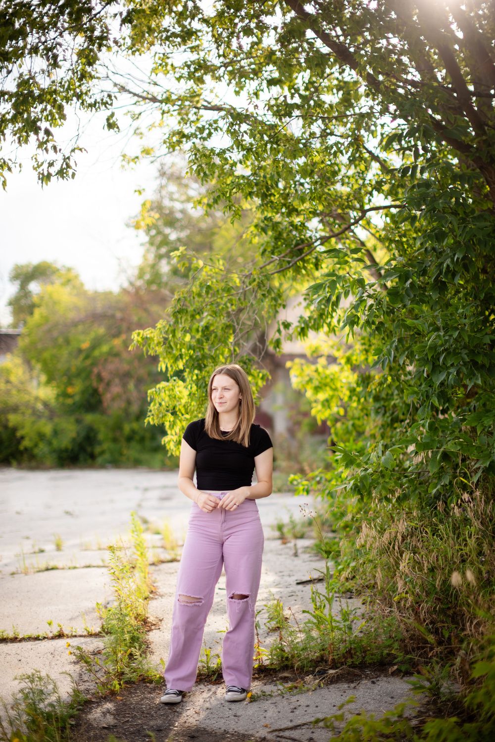 A figure in a black t-shirt and lilac ripped jeans poses outdoors among green foliage in golden sunlight.