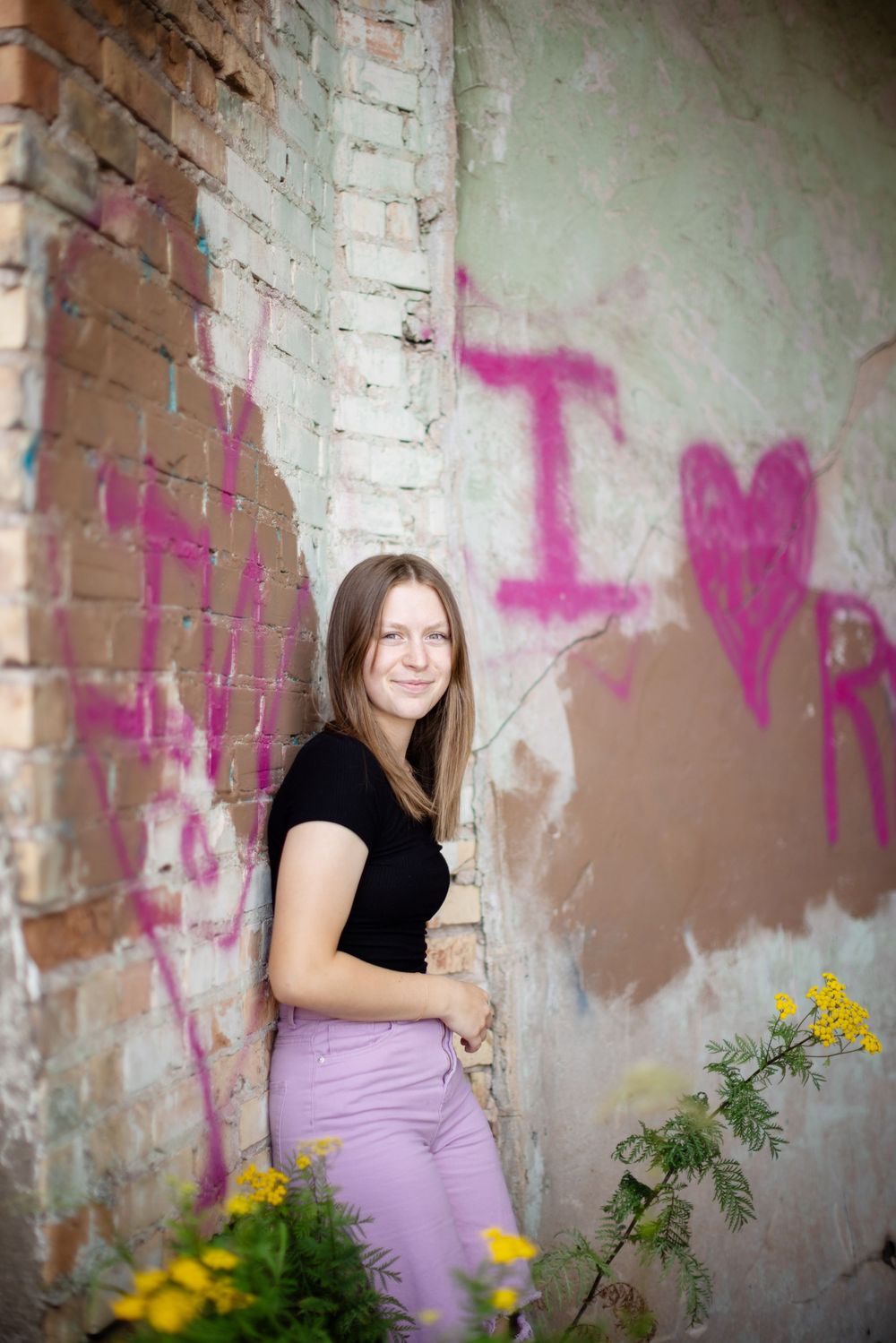 Person in a black shirt and purple pants poses by a brick wall with graffiti and yellow wildflowers in the foreground.