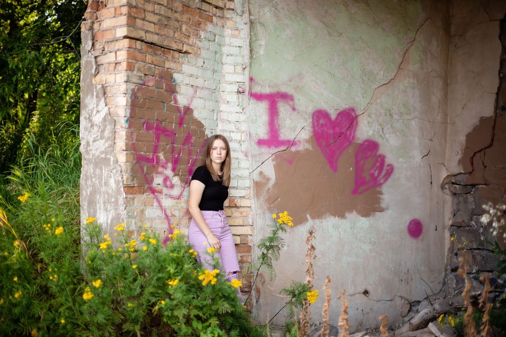 Someone sits against a concrete wall with pink graffiti hearts while yellow wildflowers bloom in the foreground.