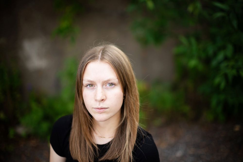 A person with long brown straight hair poses outdoors against a blurred green foliage background.