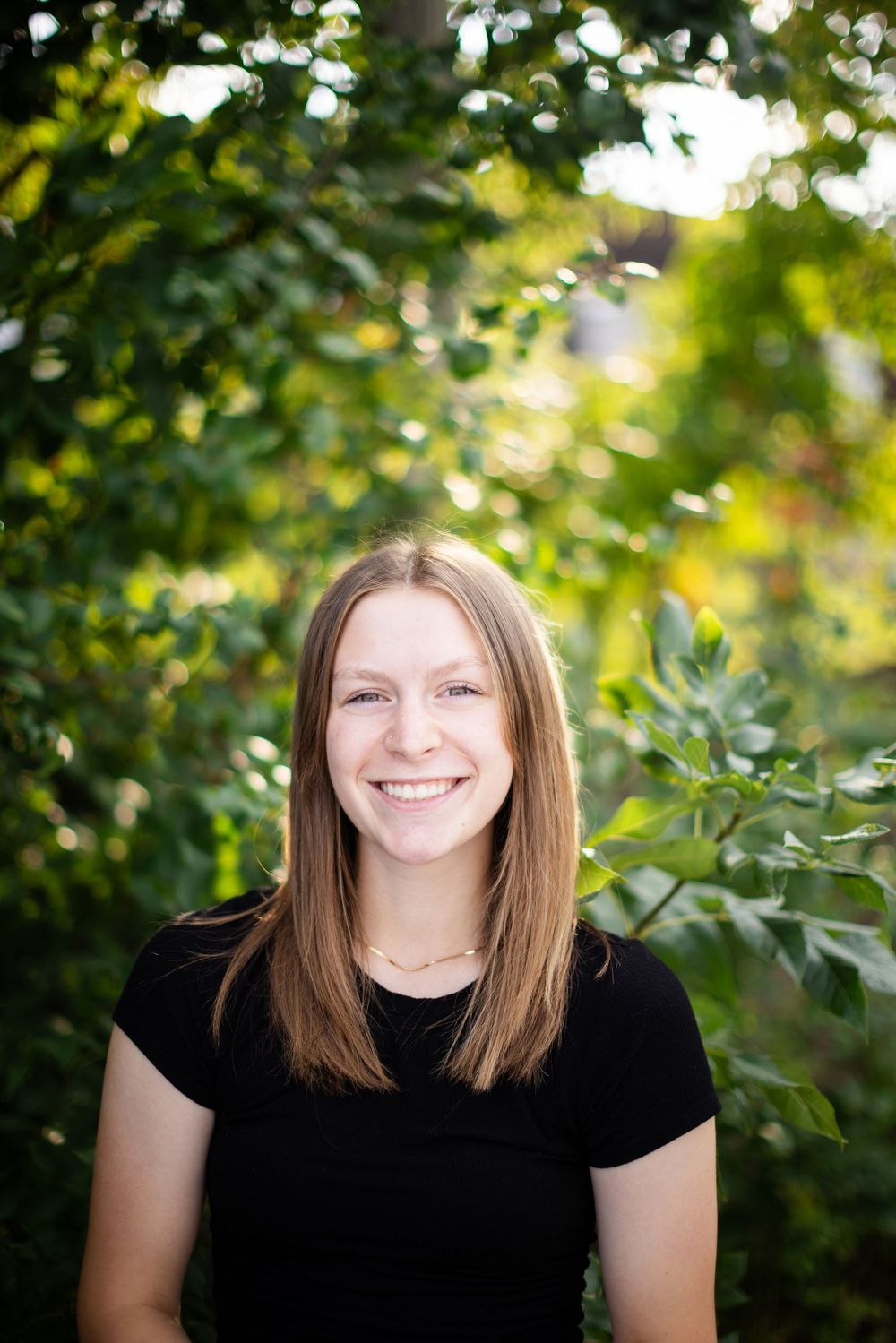 Portrait of someone in a black t-shirt smiling outdoors with green foliage and natural bokeh in the background.