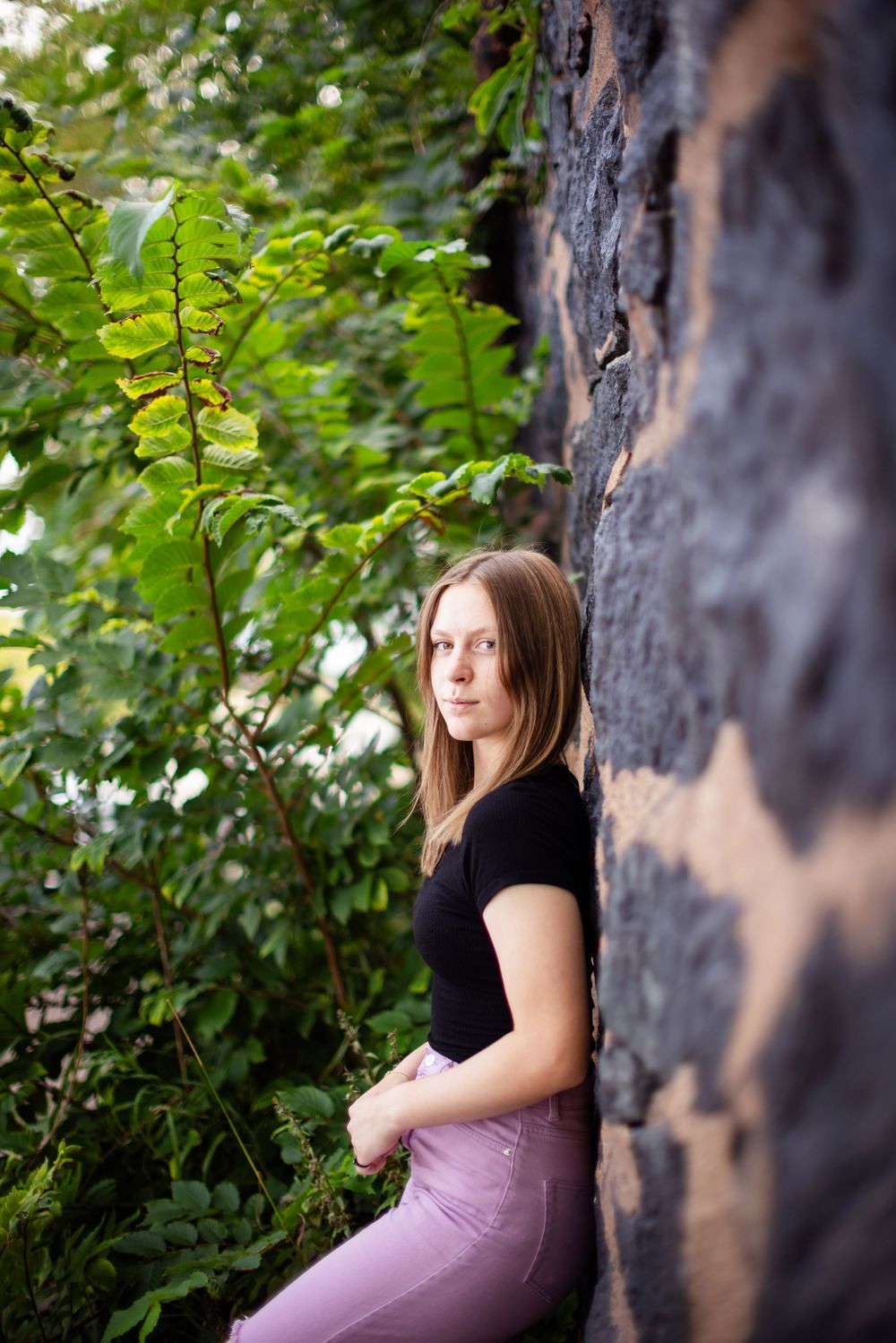 Young person in black top poses against dark textured wall surrounded by green foliage in nature setting.